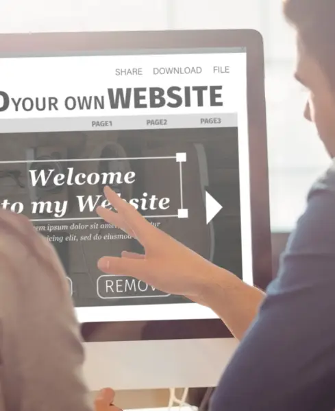 A person points at a computer screen displaying a website editor with "Welcome to my Website" text and navigation buttons.